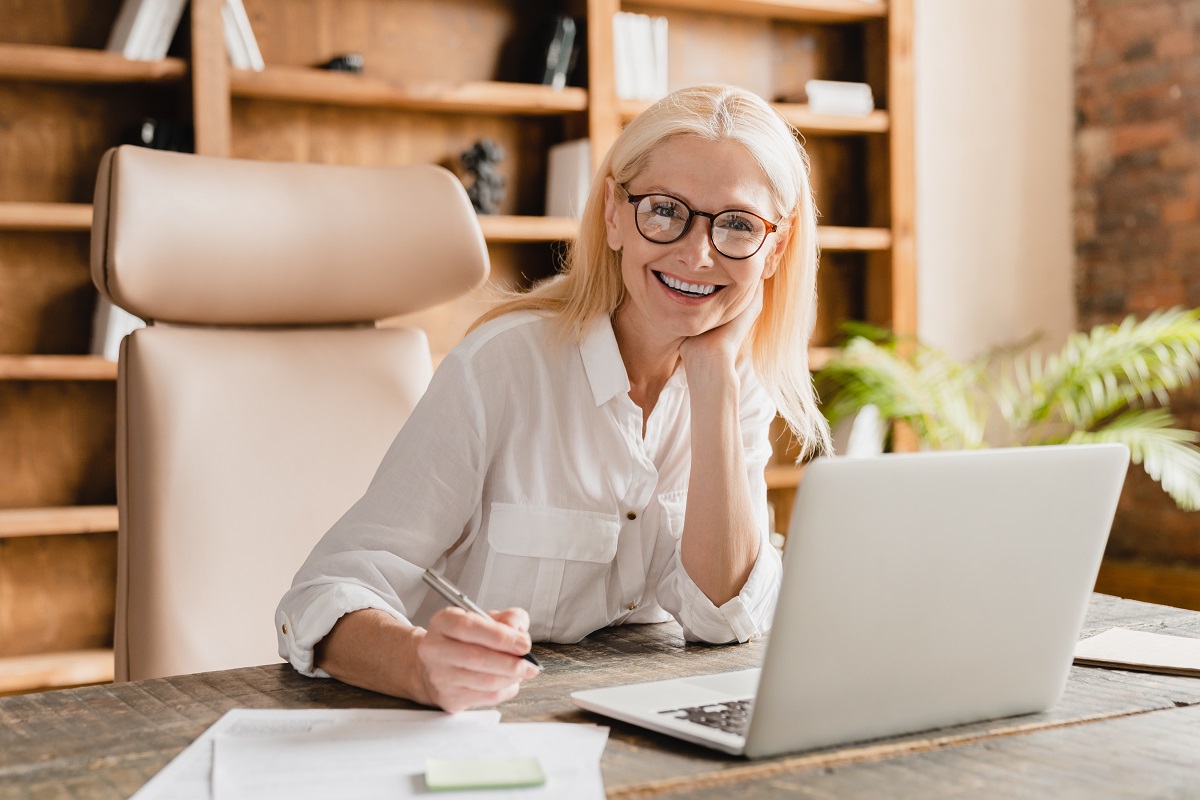Happy smiling mature middle-aged businesswoman tutor boss ceo freelancer teacher doing paperwork, signing contracts, documents while working on laptop in office
