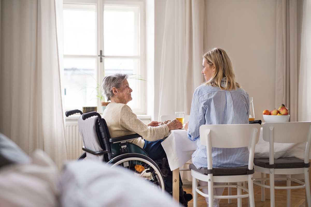 A senior woman in wheelchair with a health visitor sitting at the table at home, eating.
